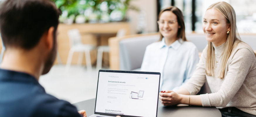 Drei Personen sitzen an einem Tisch und besprechen sich. Wir schauen einem Mann über die Schulter. Er hat einen Laptop vor sich und nutzt Secure Password Share.