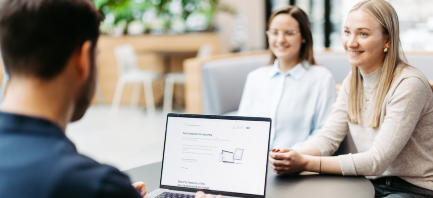 Drei Personen sitzen an einem Tisch und besprechen sich. Wir schauen einem Mann über die Schulter. Er hat einen Laptop vor sich und nutzt Secure Password Share.