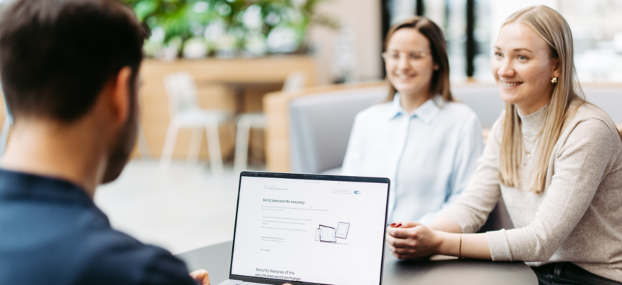 Drei Personen sitzen an einem Tisch und besprechen sich. Wir schauen einem Mann über die Schulter. Er hat einen Laptop vor sich und nutzt Secure Password Share.