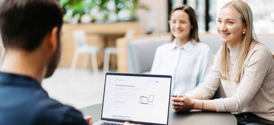 Drei Personen sitzen an einem Tisch und besprechen sich. Wir schauen einem Mann über die Schulter. Er hat einen Laptop vor sich und nutzt Secure Password Share.