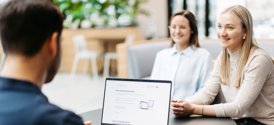 Drei Personen sitzen an einem Tisch und besprechen sich. Wir schauen einem Mann über die Schulter. Er hat einen Laptop vor sich und nutzt Secure Password Share.