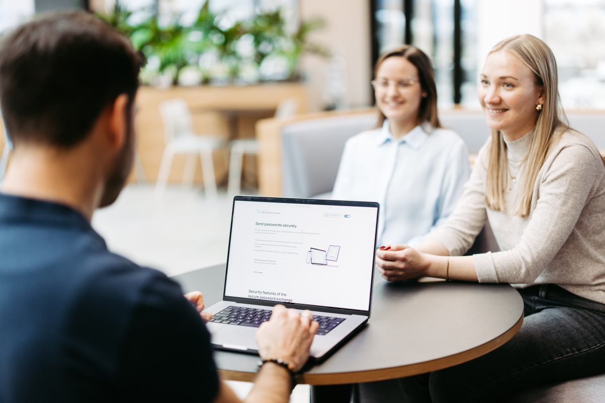 Three people are sitting at a table discussing something. We look over a man's shoulder. He has a laptop in front of him and is using Secure Password Share.